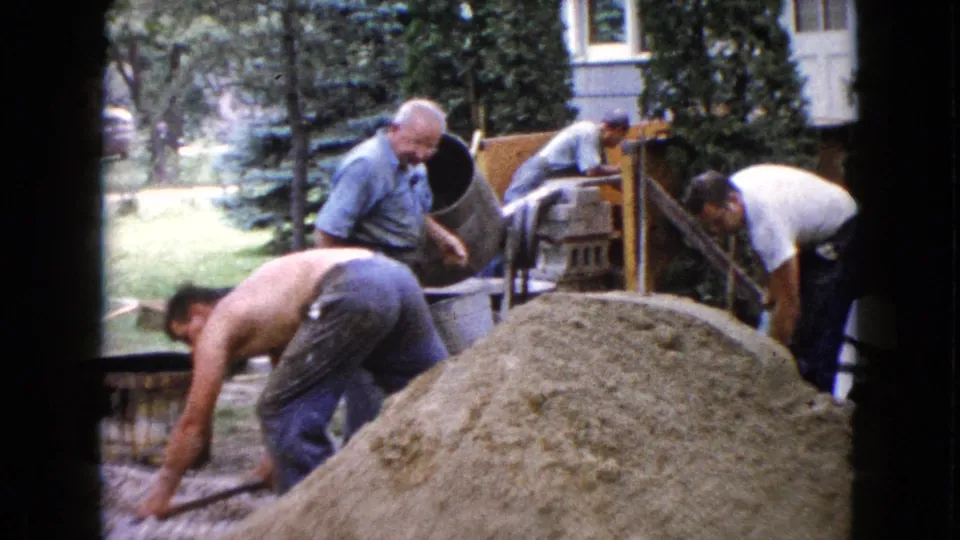 1960 Wauconda home-movie frame with men bending and shoveling near a sand pile, cement mixer, and house-side yard.