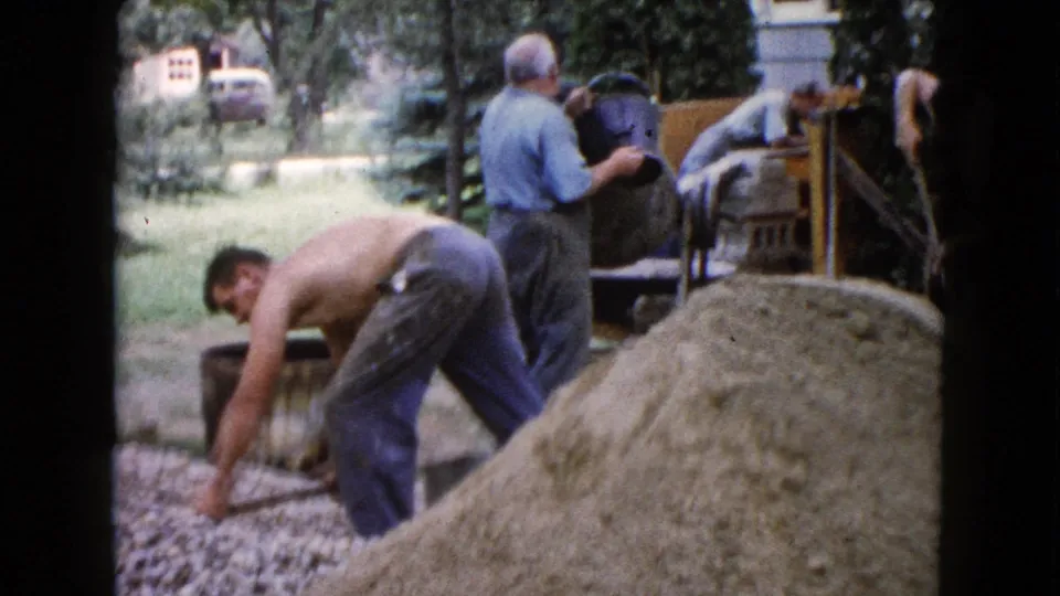 A 1960 Wauconda home-movie frame shows one man bending over gravel while another carries a dark container by the mixer.
