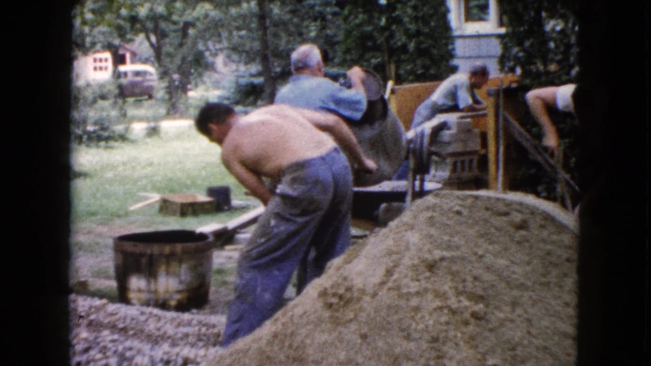 Men work around a sand pile, barrel, and concrete mixer in a residential yard.