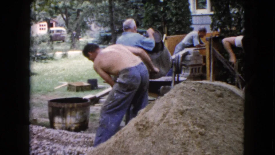 1960 Wauconda home-movie frame showing men handling construction materials beside a sand pile and mixer in a backyard.