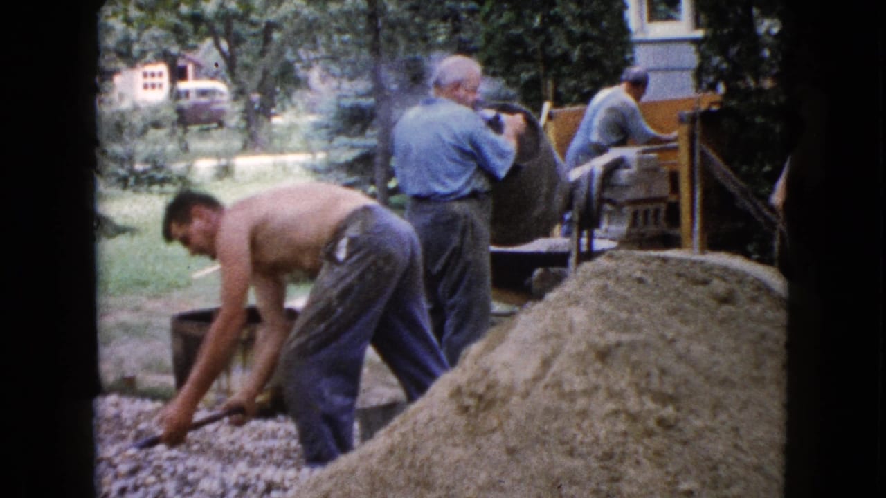 A shirtless man bends toward gravel while other men work near a concrete mixer.
