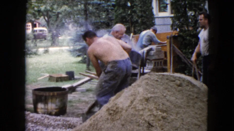 Men continue mixing concrete materials in a backyard work area, with smoke or dust near the mixer and a house behind them.