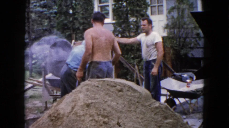 A backyard construction scene in Wauconda, 1960, with men beside a mixer, sand pile, wheelbarrow, and house steps.