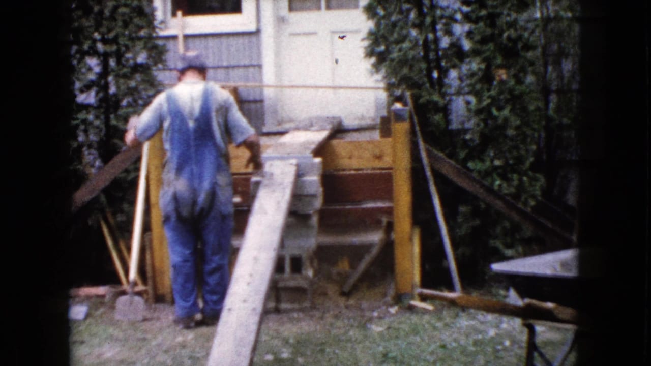 Man in blue overalls stands near a wooden ramp, concrete blocks, and porch framing in a backyard.