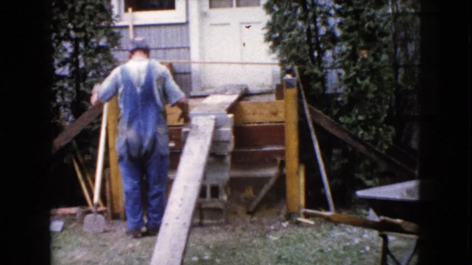 In Wauconda, Illinois, a man in blue overalls works beside a wooden ramp, concrete blocks, and porch framing in 1960 home-movie footage.