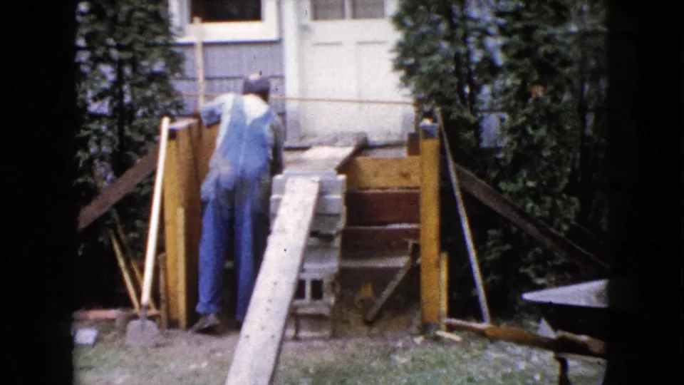 A man in work overalls leans toward porch materials near a wooden ramp and concrete blocks in the Wauconda backyard scene.