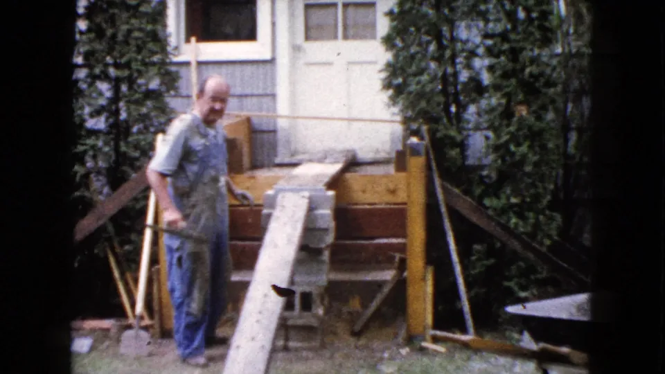 The builder turns toward the camera near the ramp and concrete blocks, with the house exterior and porch work visible behind him in Wauconda.