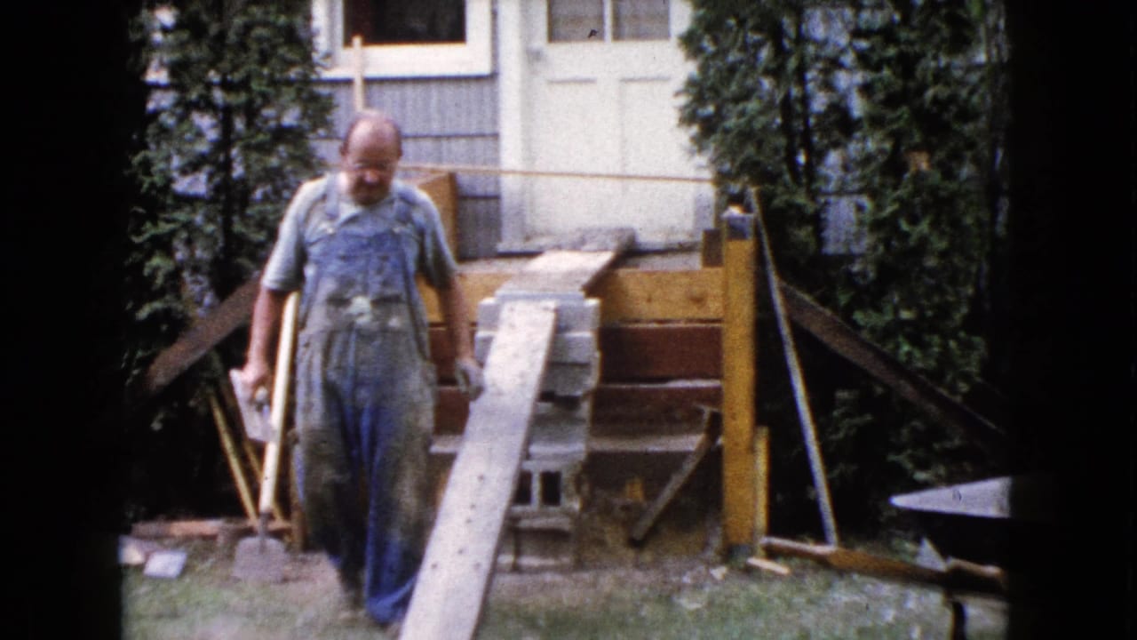 Man in blue overalls walks near a wooden plank and cinder blocks beside a house.