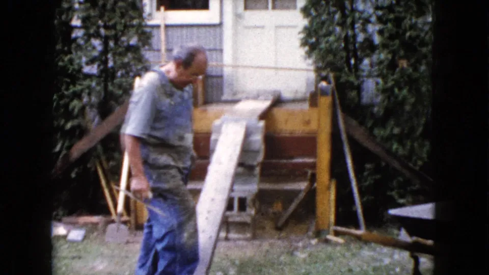 A worker in blue overalls passes the wooden ramp and unfinished deck framing in a 1960 Wauconda backyard scene.