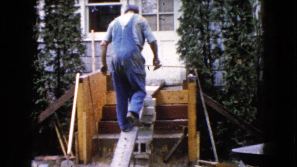 The worker climbs the plank ramp toward the unfinished deck, with cinder blocks, rail framing, shrubs, and the house exterior behind him.