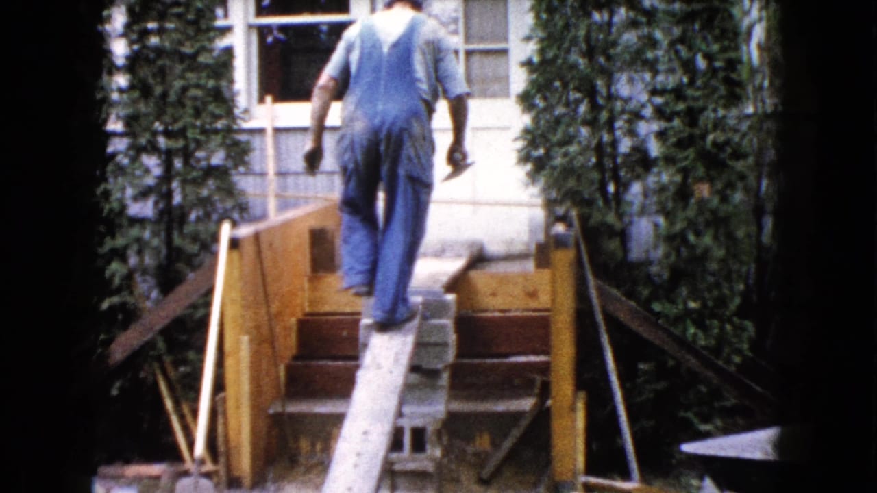 Man in blue overalls walks up unfinished wooden backyard stairs beside a plank ramp.