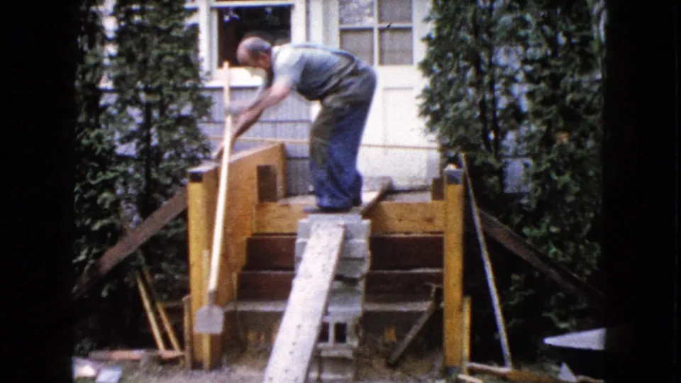 The older man extends an arm along the stair rail, likely checking placement or measurement during backyard construction work.