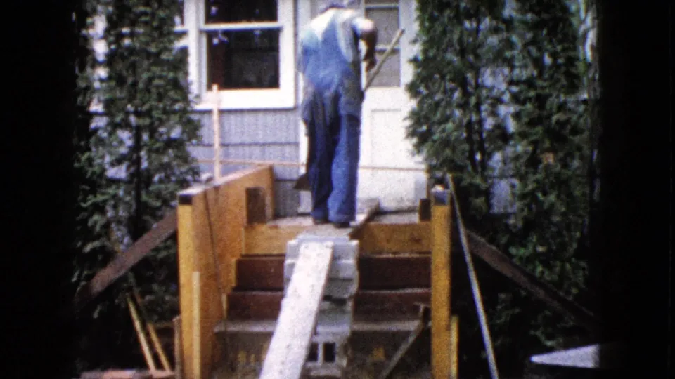 The man stands on the upper steps with his back to the camera, framed by the house exterior and tall greenery in 8mm footage.
