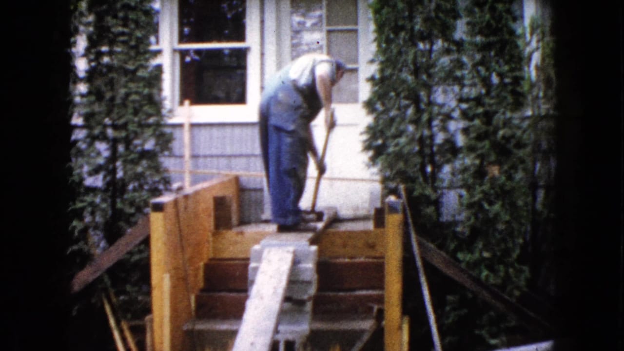 Man in blue overalls bends over porch steps with a hand tool near a house entrance.