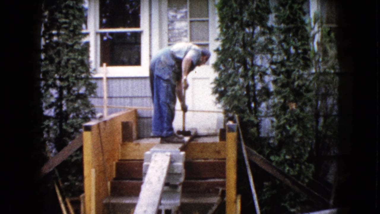 The 1960 home-movie frame shows a man continuing porch work outside a Wauconda house, with wooden rails, steps, siding, and tall shrubs visible.