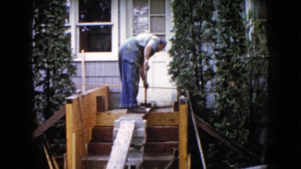The 1960 home-movie frame shows a man continuing porch work outside a Wauconda house, with wooden rails, steps, siding, and tall shrubs visible.