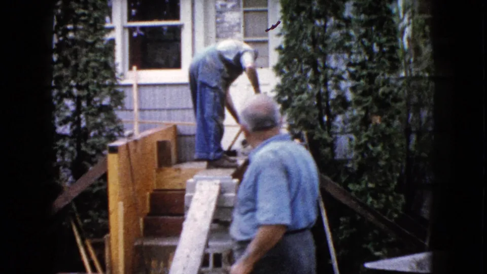 A second adult man appears in the foreground while the worker in overalls continues repairing or building the porch steps outside the Wauconda house.