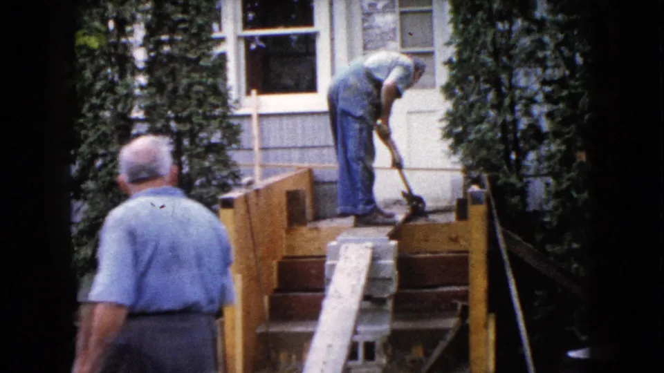 The foreground observer stands with his back to the camera while the man in overalls works on the porch steps in this 1960 Wauconda home-movie frame.