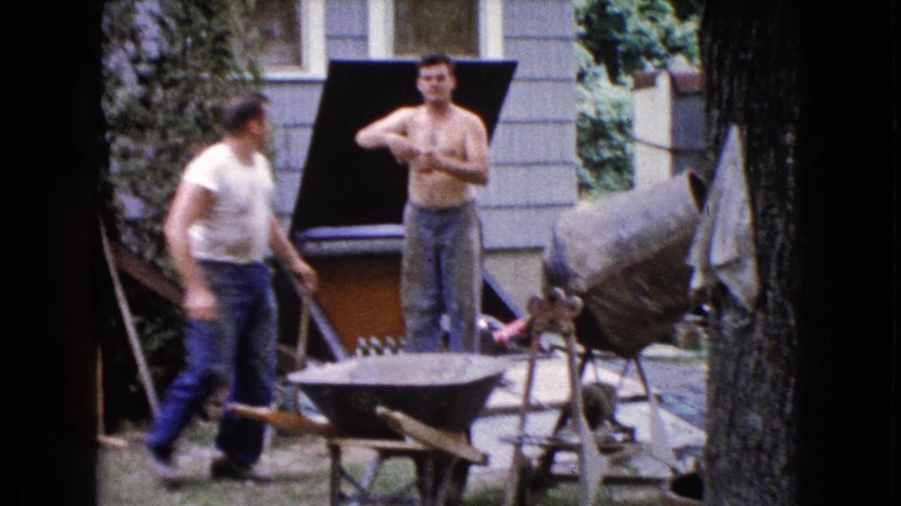 Two adult men in a backyard near a cement mixer, wheelbarrow, house siding, and wooden steps.