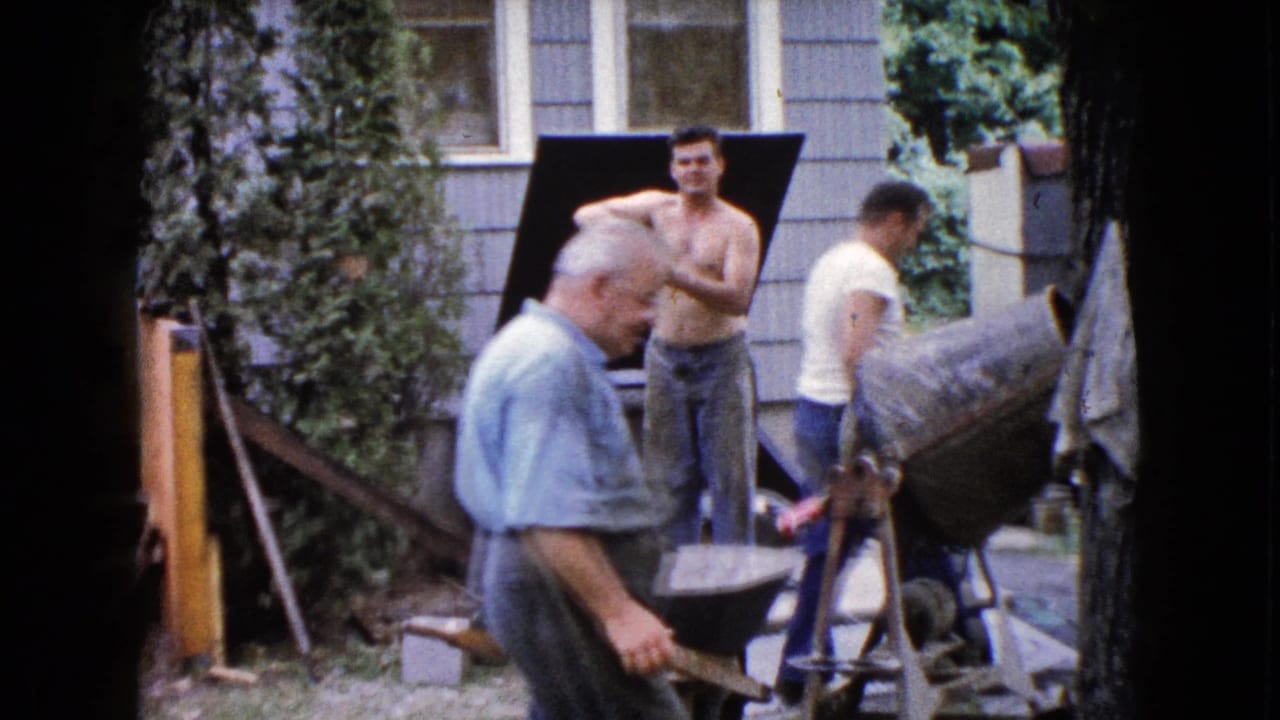 Older man walks in front of a cement mixer while two other men stand near the house.