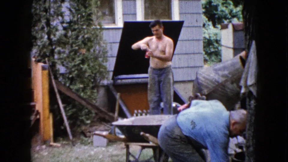 The shirtless man remains by the house and cement mixer as an older man bends down near the wheelbarrow in the backyard work area.