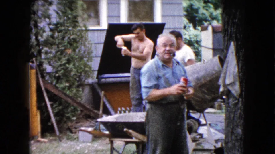 In the Wauconda backyard, an older man in a blue shirt smiles at the camera with a red cup while work tools and two men remain behind him.