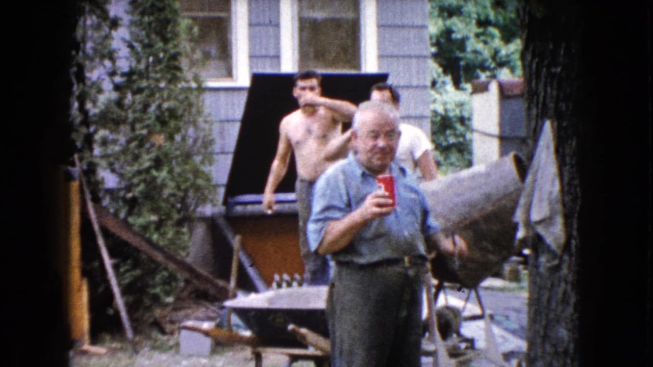 Older man drinks from a red cup as a shirtless younger man stands behind him near a grill.
