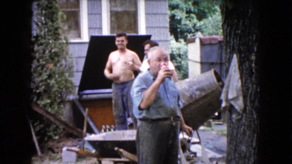 In the 1960 Wauconda home-movie scene, the older man drinks in the foreground as younger men linger near a grill and yard equipment.