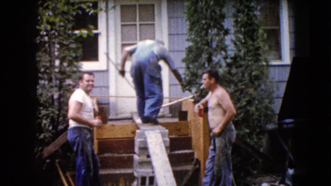 A 1960 Wauconda home-movie frame shows one man bending on porch steps while two other men watch near a plank and shrubs.