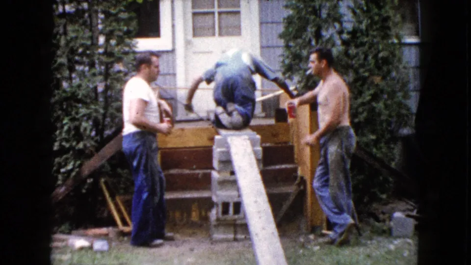 A Wauconda, Illinois 1960 home-movie frame shows one man crouched on the porch steps while two men stand beside the work area.