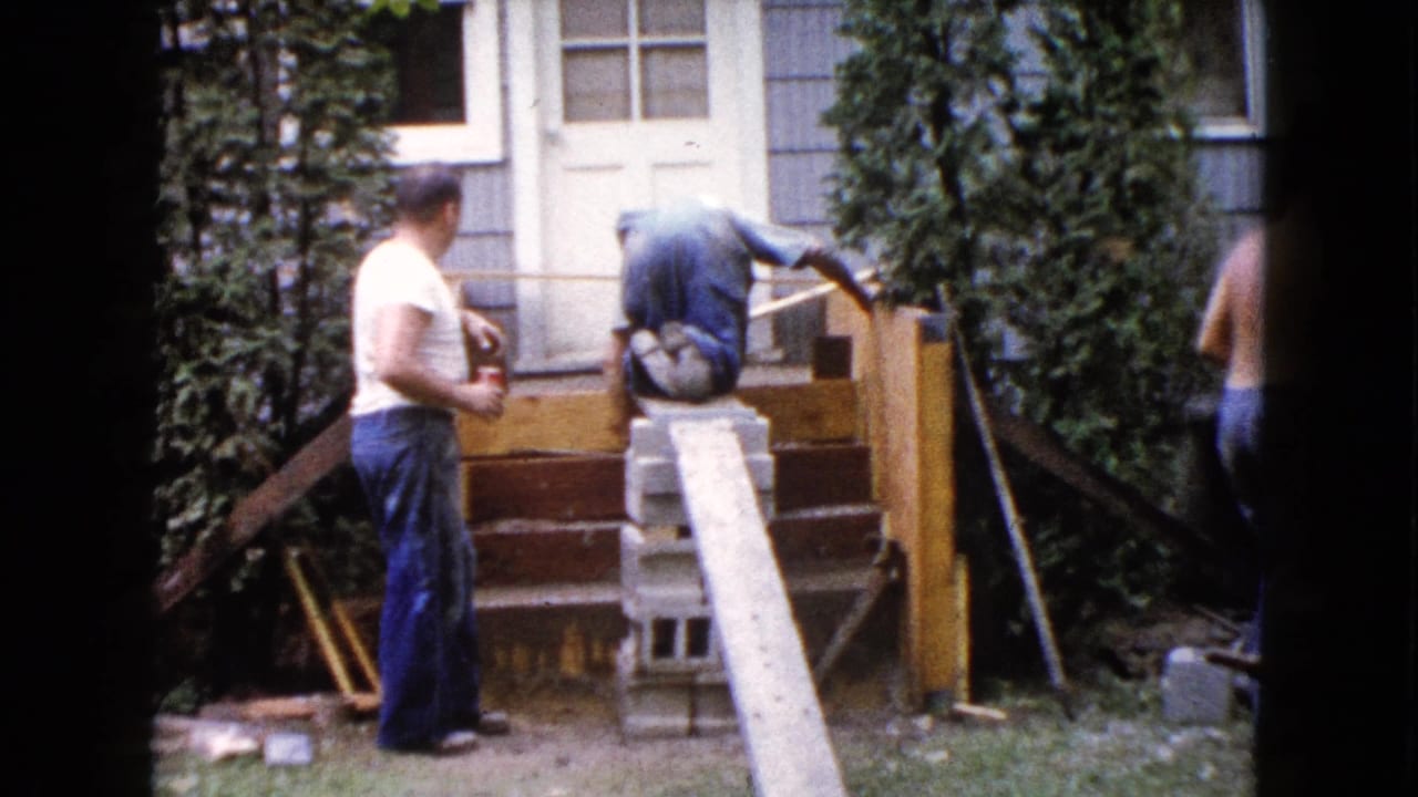 A man in a white t-shirt watches as another bends over the wooden steps, with a shirtless man partly leaving the frame in the 1960 Wauconda backyard scene.