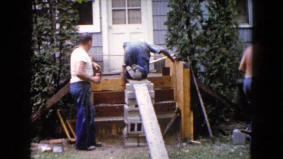 A man in a white t-shirt watches as another bends over the wooden steps, with a shirtless man partly leaving the frame in the 1960 Wauconda backyard scene.