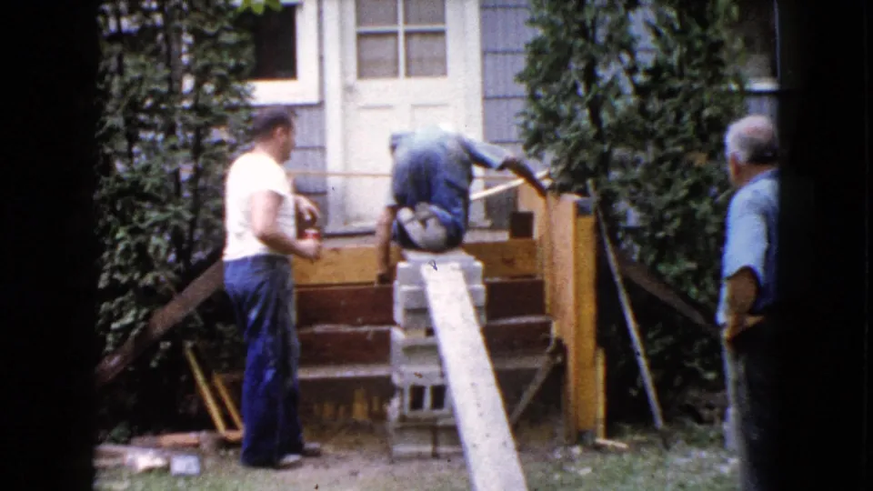 The 1960 home-movie frame shows a man in a white t-shirt near the steps while another adult bends over the wooden structure beside shrubs and house siding.
