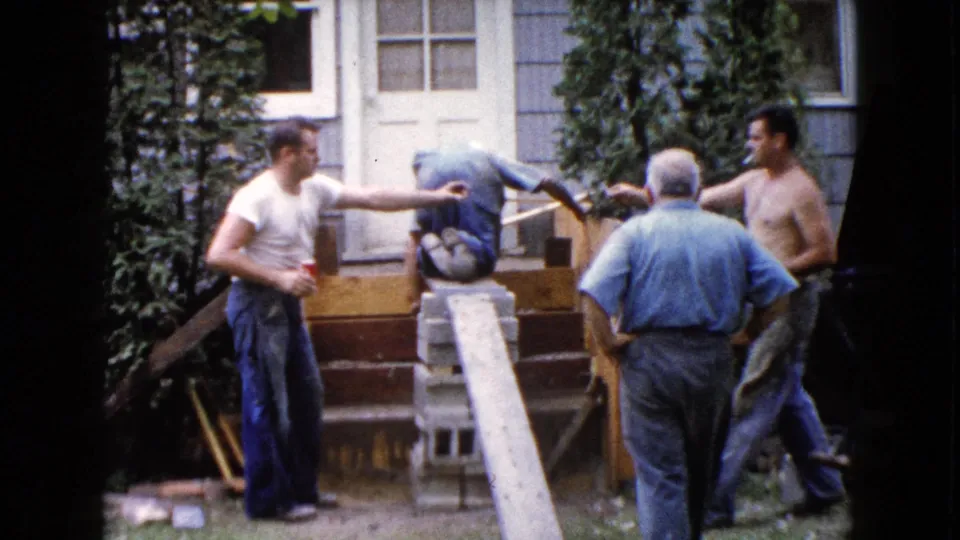 Three adult men confer around the wooden backyard steps in Wauconda, Illinois, with one pointing as the 8mm frame records the home improvement scene.