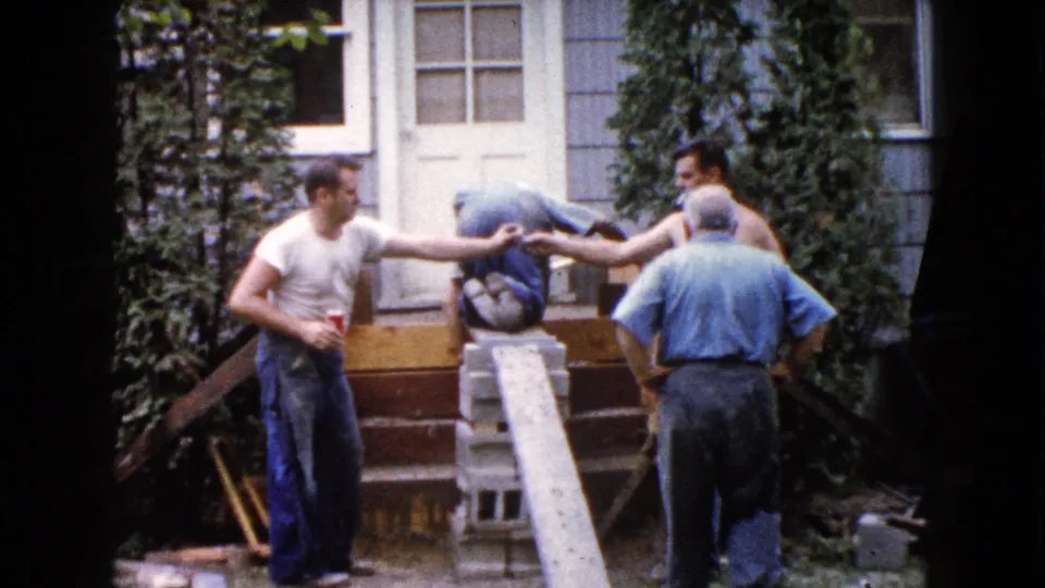 In Wauconda, Illinois, 1960, four adult men work outside a light-sided house with a plank, blocks, and hand tools near the front steps.