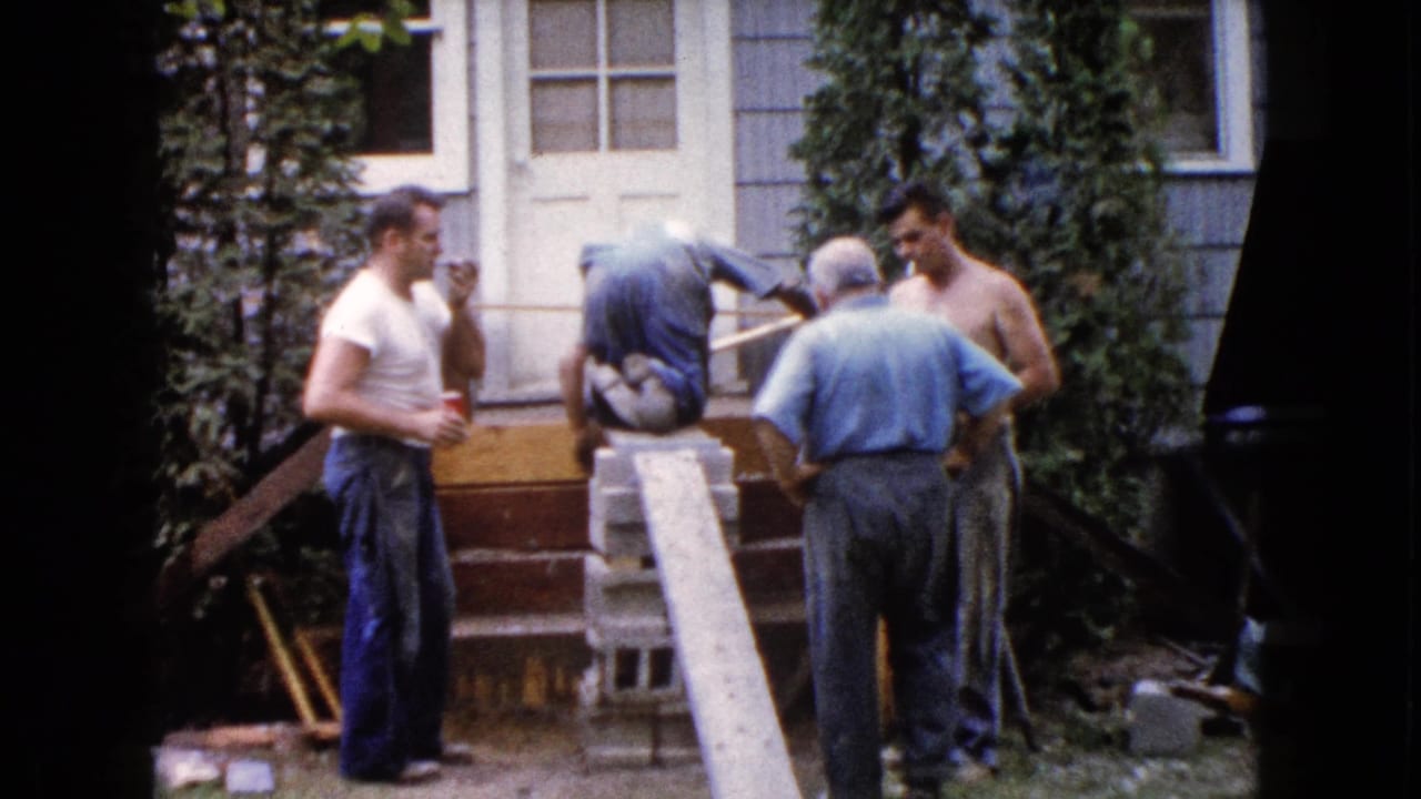 Men stand and kneel near a house doorway with a plank, blocks, and tools in the yard.