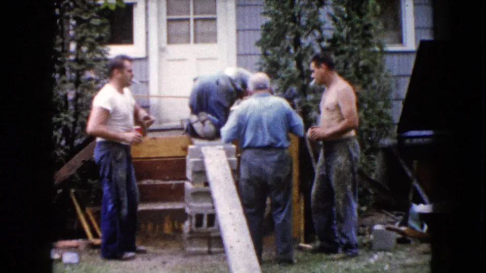 A 1960 home-movie frame shows men at a Wauconda house renovation, gathered around the front steps with blocks, a long plank, and tools.