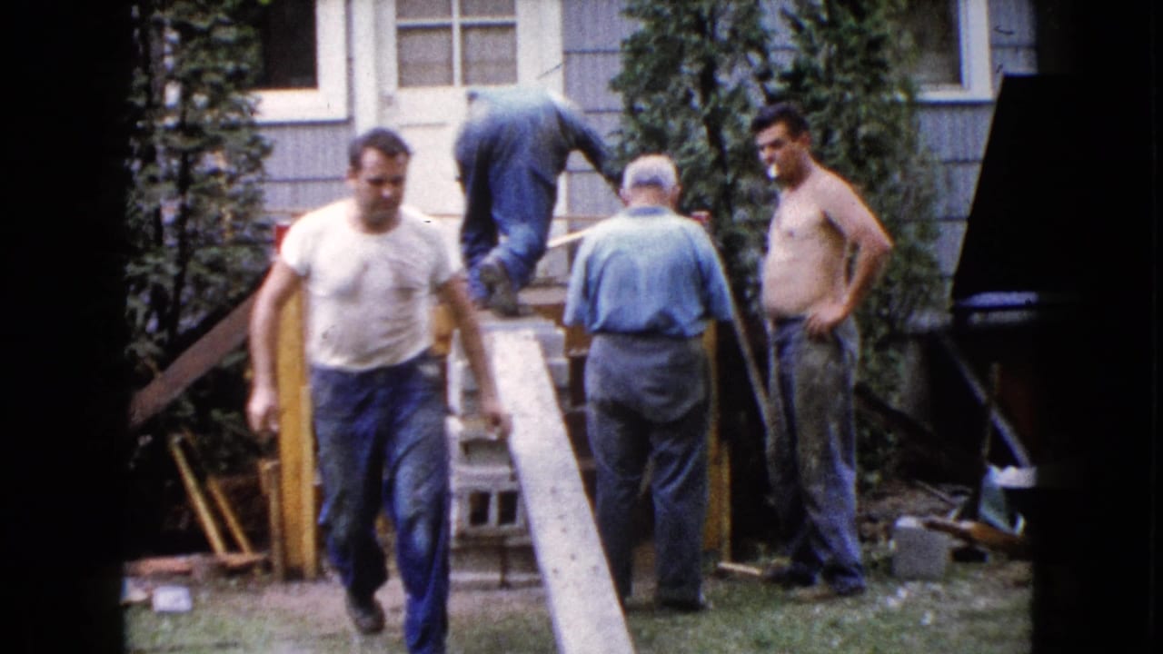A 1960 Wauconda home-movie frame shows a man in a white shirt walking away from deck work outside a house.