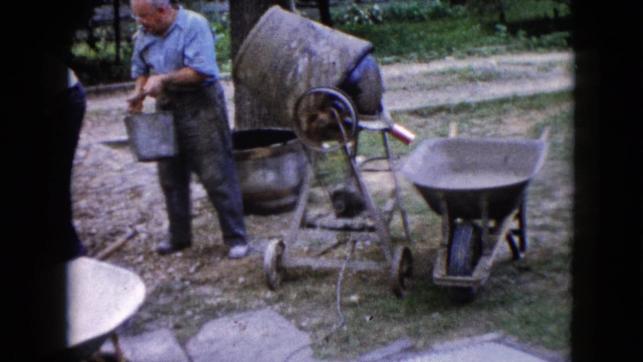 An adult man works beside a portable cement mixer in a Wauconda backyard, with buckets, gravel, and a wheelbarrow nearby.