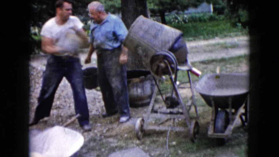 Two adult men handle buckets near a portable cement mixer and wheelbarrow during a 1960 backyard concrete job in Wauconda.