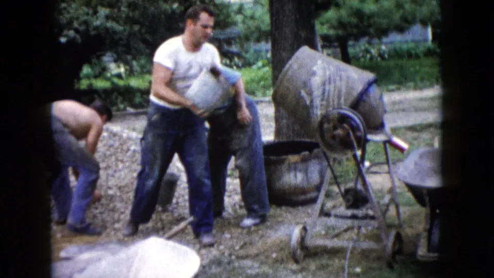 An adult man carries a bucket in front of a cement mixer while another man bends near the work area in the Wauconda backyard scene.