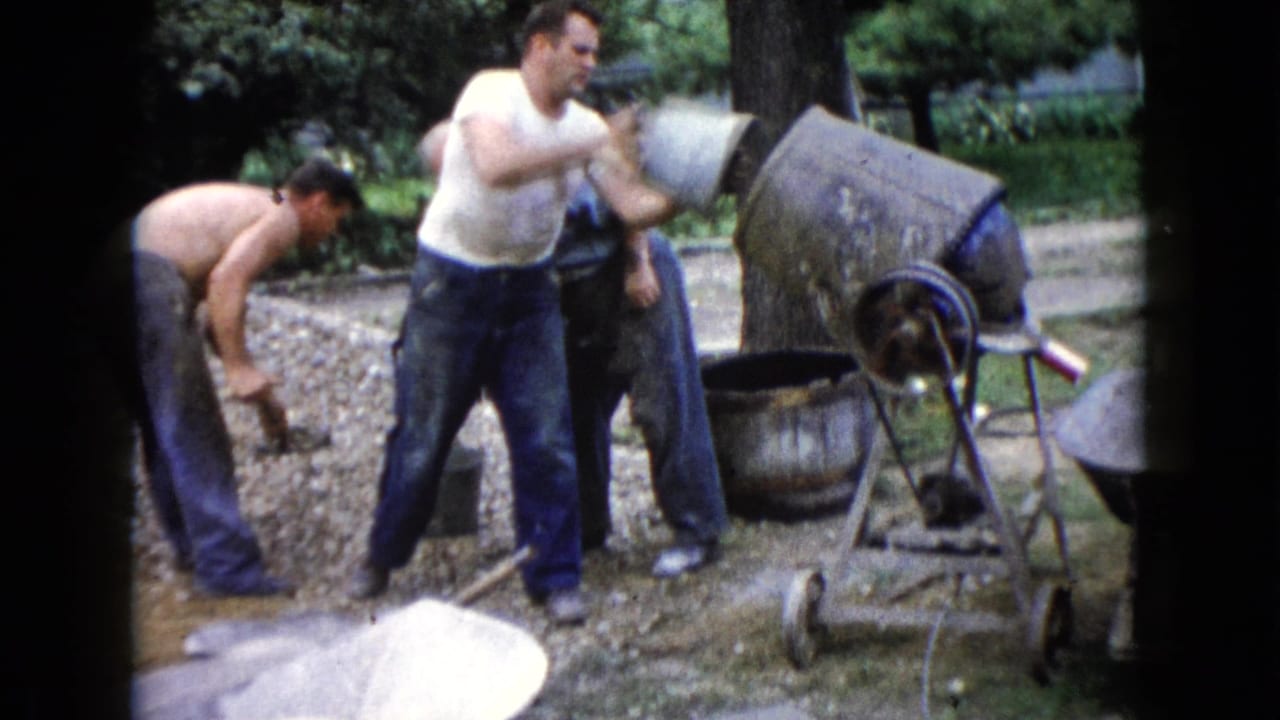 Three adult men work near a gravel pile, bucket, wheelbarrow, and concrete mixer outdoors.
