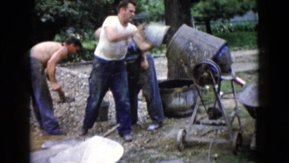 In 1960 Wauconda, Illinois, three adult men appear to shovel and pour material beside a gravel pile and concrete mixer.