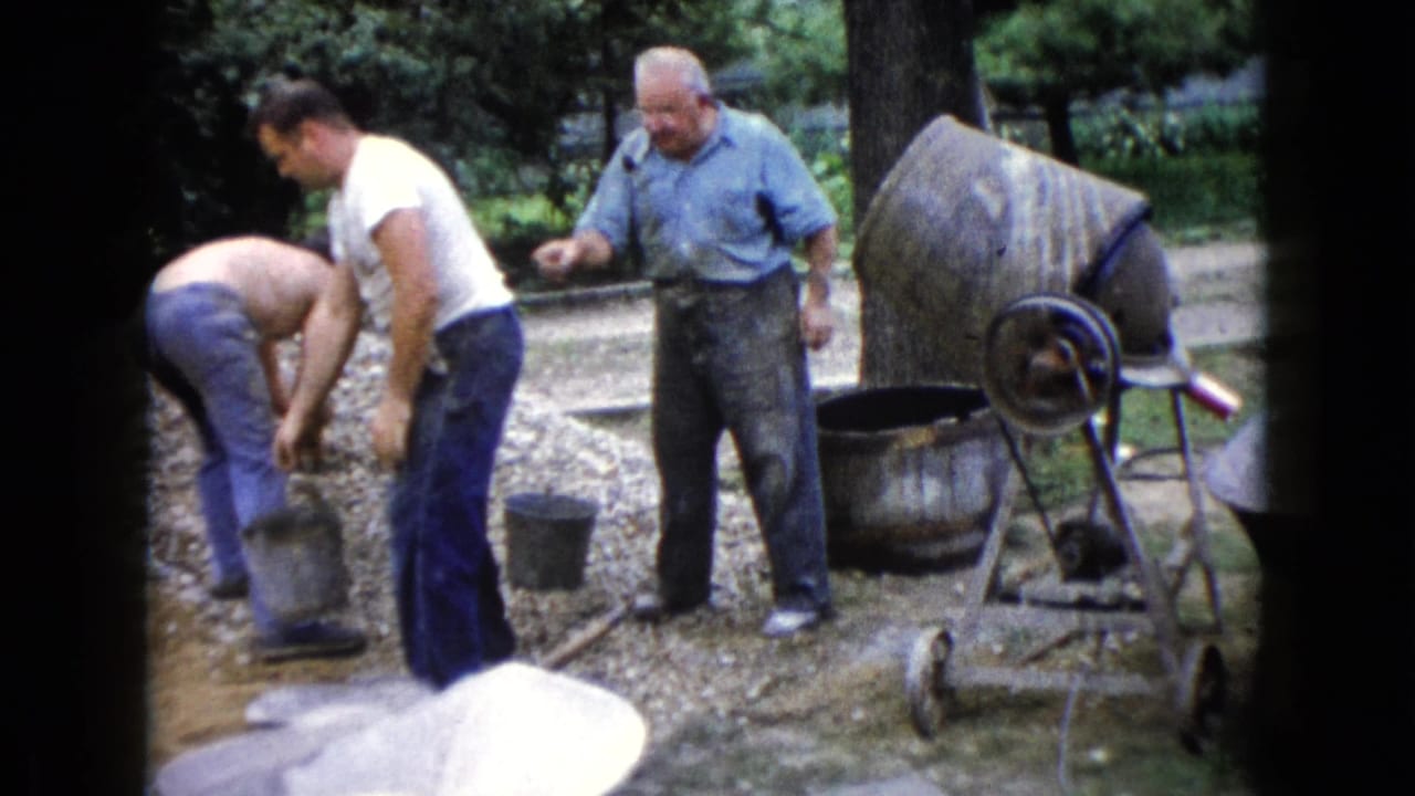 Two men handle a bucket near a gravel pile while a third man stands beside a concrete mixer.