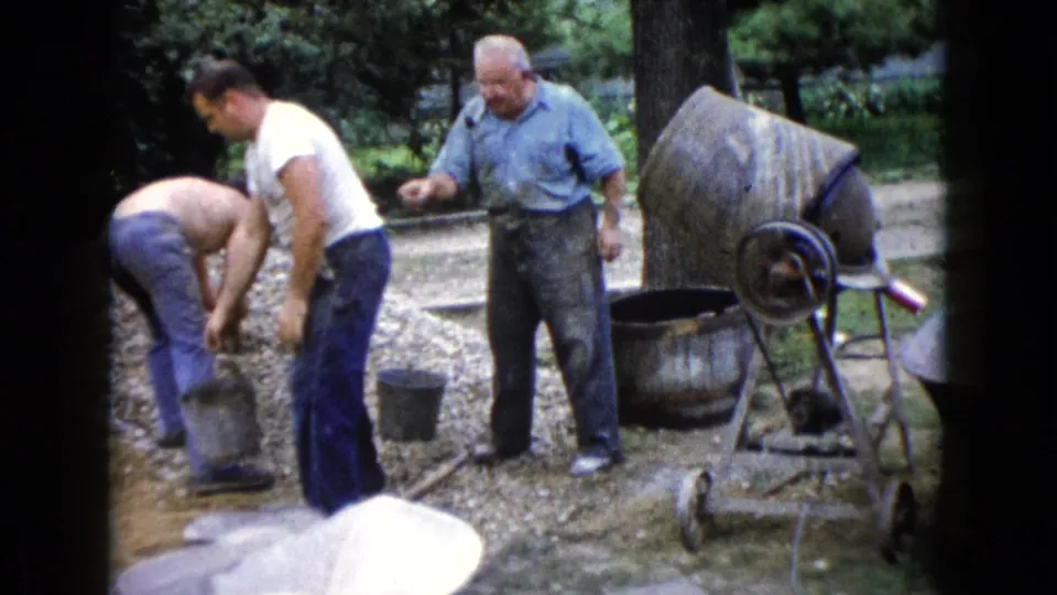 In the Wauconda home-movie scene, two men handle a bucket and gravel while another man stands near the mixer.