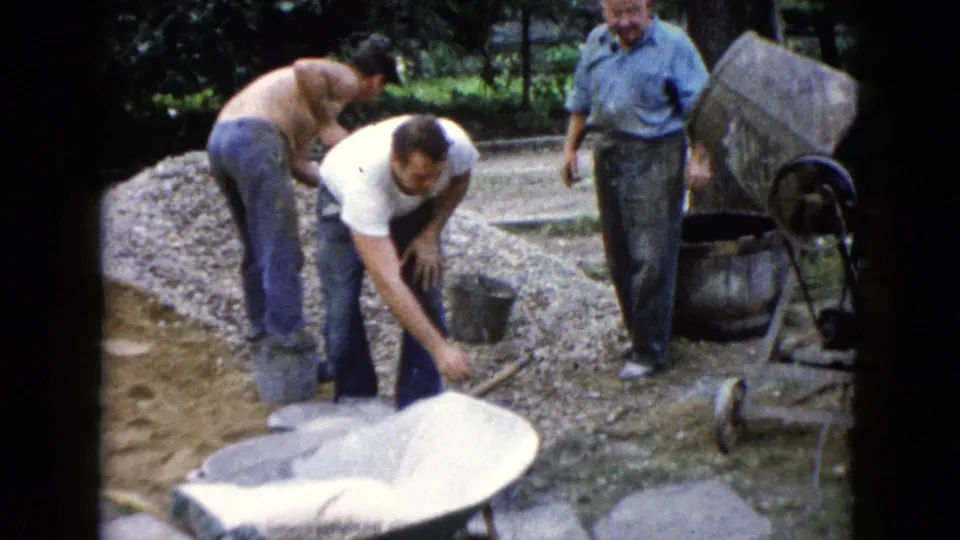 A 1960 Illinois home-movie frame shows men working around a gravel pile, wheelbarrow, shovel, and concrete mixer.