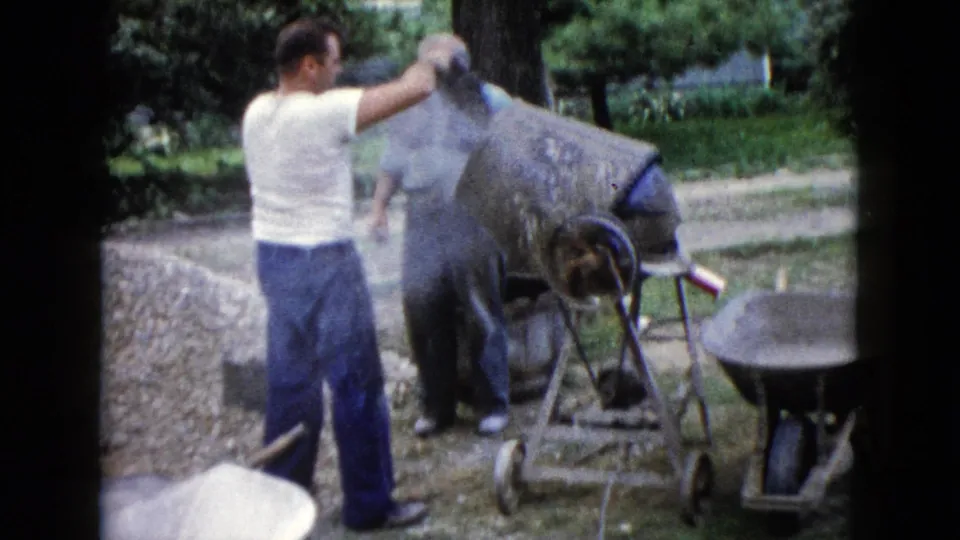 In Wauconda, Illinois, two adult men appear to work with a cement mixer near a wheelbarrow and gravel pile in 1960 home-movie footage.