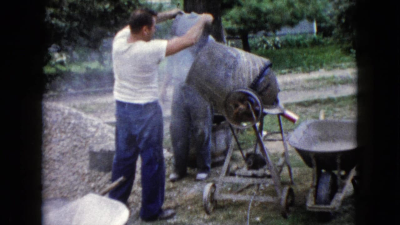 A man in a white shirt works at the cement mixer as another adult stands nearby in a residential yard in Wauconda, Illinois.