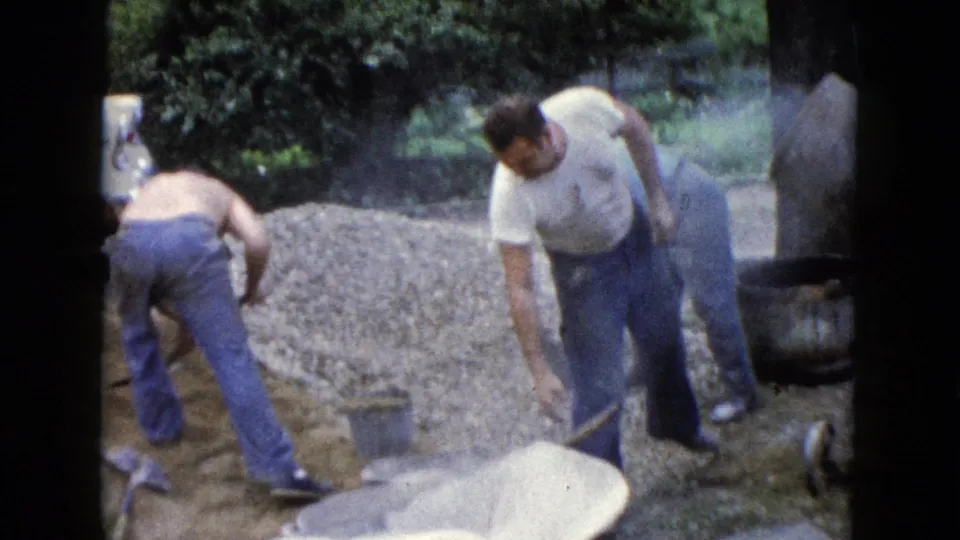 Two adult men appear to shovel or gather material near a gravel pile in a Wauconda yard, with the mixer and wheelbarrow partly visible.