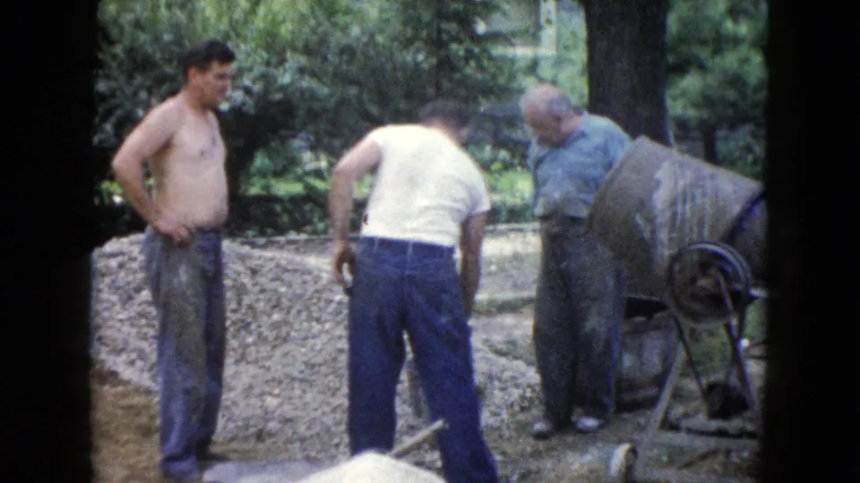 In this 1960 Wauconda home-movie frame, men in work clothes stand around a cement mixer beside gravel and yard tools.